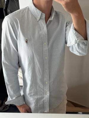 Blåvit randig skjorta med knappar - Säljer en klassisk blåvit randig skjorta med button-down krage och långa ärmar. Skjortan har knappar hela vägen och en liten broderad logga på bröstet. Perfekt till jeans eller chinos för en clean och stilren look.