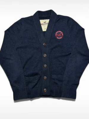 Hollister button up cardigan - Mörkblå kofta från Hollister med knappar framtill och två fickor. Storlek L men passar som M. Klassisk design med ribbade muddar och ett rött Hollister-märke på bröstet. Perfekt för lager på lager och enkel att matcha med olika outfits. Mått: 64 cm lång 51 cm bred. Säljes då jag själv är 187 och brukar ha L, men den var för liten. 