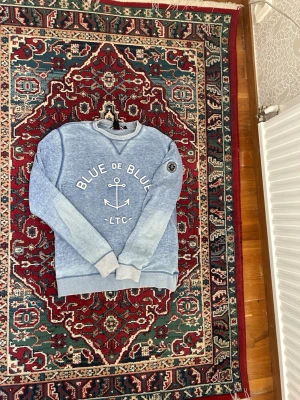 Blå sweatshirt Le Temps des Cerises - Blå sweatshirt från Le Temps des Cerises med tryck 'BLUE DE BLUE' och ett ankare på bröstet. Tröjan har rund halsringning, ribbade muddar och en avslappnad passform. Perfekt för en casual look och tillverkad i mjukt material.