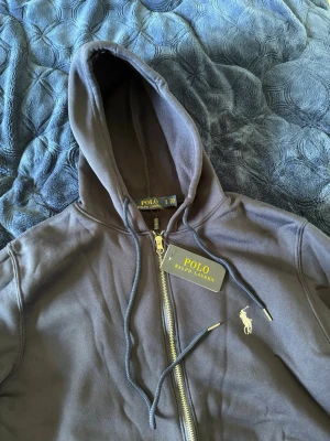 Svart hoodie från Polo Ralph Lauren - Snygg svart hoodie från Polo Ralph Lauren med dragkedja framtill och klassisk huva. Ikoniska Polo-loggan broderad i vitt på bröstet. Tillverkad i mjukt bomullsmaterial och har justerbara snören i huvan. Perfekt för en chill och stilren look.