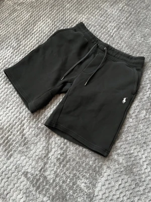 Svarta shorts med snörning från Polo - Svarta shorts från Polo Ralph Lauren med klassisk logga broderad på vänster ben. Mjukt material, elastisk midja med snörning och två sidofickor. Perfekta för en avslappnad stil och passar till många tillfällen. Passar dig som har Small i storlek då dom krympt i tvätten!!!