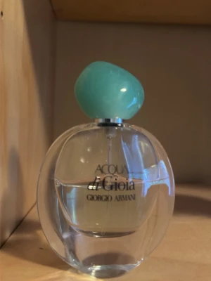 Acqua di Gioia parfym Giorgio Armani - Acqua di Gioia parfym från Giorgio Armani i en rund, transparent glasflaska med turkosgrön, stenliknande kork. Flaskan har elegant svart text och en stilren design som känns både fräsch och modern. Perfekt för dig som gillar exklusiva dofter.