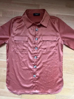 Rostfärgad satinblus med knappar Sisters Point - Snygg rostfärgad skjorta i glansigt satinmaterial med klassisk krage och knappar framtill. Skjortan har två bröstfickor med lock och halvlånga ärmar. Perfekt för dig som vill ha en trendig och stilren look.
