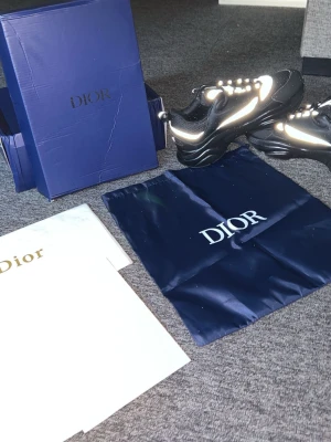 Svarta Dior sneakers med vita detaljer - Säljer ett par svarta Dior sneakers med vita och reflekterande detaljer. Skorna har en chunky sula, snörning och en modern, sportig look. Kommer med originalkartong, dustbag och kvitto. Perfekta för dig som vill ha något exklusivt och trendigt.