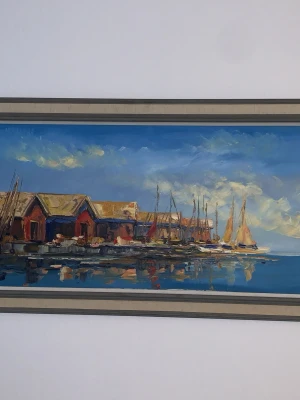 Oljemålning med båthus och segelbåtar 114x60 cm - Säljer en färgstark oljemålning i landskapsformat som föreställer röda båthus och segelbåtar vid en brygga. Tavlan har en bred ram i beige och grå toner. Motivet har tydliga penseldrag och en blå himmel med moln reflekteras i vattnet.