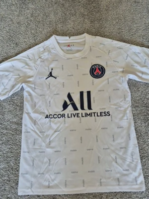 Vit PSG tröja Jordan x Paris - Säljer en vit PSG tröja från Jordan x Paris Saint-Germain med svart logga och texten 'Accor Live Limitless' på bröstet. Tröjan har små grå textdetaljer med 'PARIS' över hela tyget och klubbmärke på vänster bröst. Perfekt för fotboll träning och vardag.