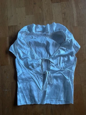 Vit glansig blus med knytning - Supersöt vit blus i glansigt material med korta ärmar och djup v-ringning. Den knyts där framme med två band. Blusen har volang vid nacken Perfekt att styla med jeans eller kjol för en trendig look.