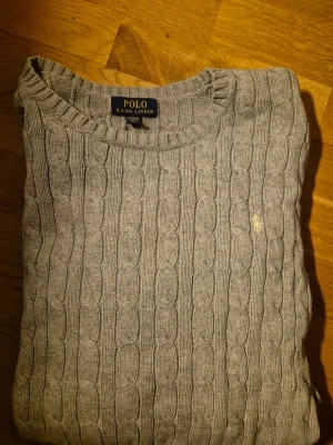Grå kabelstickad tröja Polo Ralph Lauren - Snygg grå kabelstickad tröja från Polo Ralph Lauren. Klassisk rund halsringning och diskret broderad logga på bröstet. Perfekt till jeans eller chinos för en clean och stilren look.