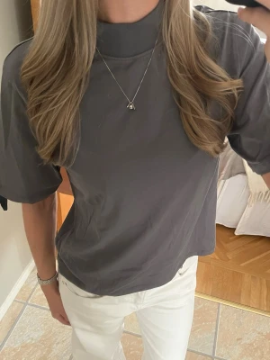 Grå t-shirt med hög hals från Lindex - Säljer en stilren grå t-shirt från Lindex med hög hals och avslappnad passform. T-shirten är tillverkad i mjuk bomull och har korta ärmar. Perfekt till jeans eller kjol för en enkel och clean look.