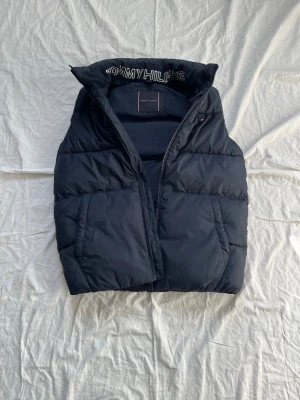 Svart dunväst från Tommy Hilfiger - Snygg svart dunväst från Tommy Hilfiger med hög krage och broderad logga på insidan av kragen. Västen har två framfickor och dragkedja framtill. Klassisk puffer-modell i mjukt och varmt material, perfekt för lager-på-lager-stil.