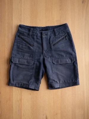 Mörkblå Sail Racing cargoshorts med dragkedjor - Snygga mörkblå cargoshorts med flera fickor, två med dragkedja framtill och två stora fickor med lock på benen. Shortsen har bälteshällor och stängs med knapp och dragkedja. Perfekta för sommaren och tillverkade i bomull för en skön känsla.