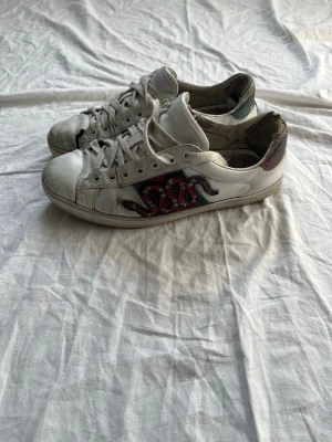 Vita sneakers med orm-motiv - Säljer ett par vita sneakers med coolt orm-motiv i rött och svart på sidan. Skorna har låga skaft, vita snören och detaljer i grönt och rött på hälen. Yttermaterialet är slätt läder och sulan är i gummi. Perfekta för dig som gillar statement-detaljer!