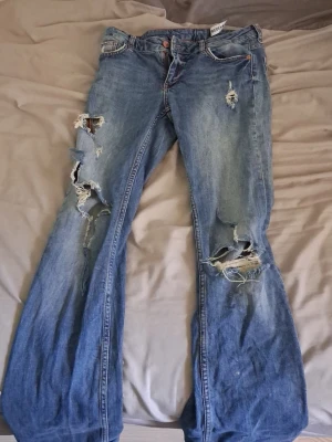 Blå slitna bootcut jeans - Säljer ett par blå bootcut jeans med rejält slitna och trasiga detaljer på både fram- och baksida. Jeansen har klassisk femficksmodell och är gjorda i denim. Perfekta för dig som gillar en edgy och avslappnad stil. Aldrig använda då dem är för långa på mig. Finns ingen lapp med storlek men dem passar som en S. 