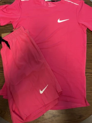Rosa träningsset  Nike miler - Säljer ett sportigt träningsset från Nike i stark rosa färg. Setet består av en t-shirt med rund hals och korta ärmar samt matchande shorts med resårmidja och snörning. Båda plaggen har vita Nike-loggor och är tillverkade i ett lätt, snabbtorkande material.
