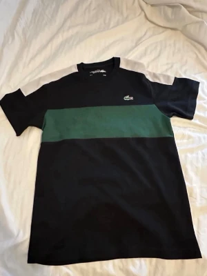 Svart t-shirt med grön rand Lacoste - Snygg svart t-shirt från Lacoste med en bred grön rand över bröstet och klassisk krokodillogga. T-shirten har rund hals och korta ärmar. Tillverkad i ett mjukt material som känns skönt mot huden.