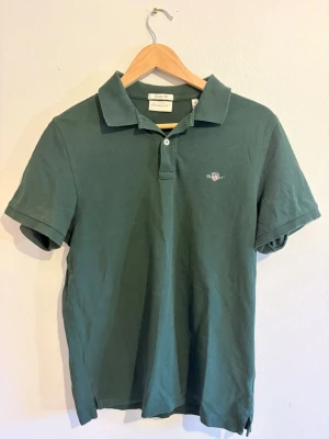 Mörkgrön pikétröja från GANT - Klassisk mörkgrön pikétröja från GANT med korta ärmar och krage. Tröjan har knappslå framtill och en liten broderad logga på bröstet. Perfekt till jeans eller shorts för en clean och stilren look.