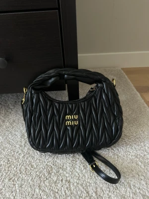 Svart handväska från Miu Miu - Snygg svart handväska från Miu Miu i quiltat läder med guldfärgade detaljer och logga framtill. Väskan har både ett kort handtag och en justerbar axelrem för olika stilar. Perfekt accessoar för att lyfta din outfit. Saknar kvitto därav priset.