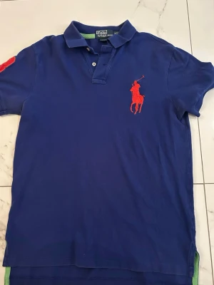 Blå pikétröja från Polo Ralph Lauren - Klassisk blå pikétröja från Polo Ralph Lauren med röd broderad logga på bröstet och krage med knappar. Tröjan har korta ärmar och är perfekt till sommaren.