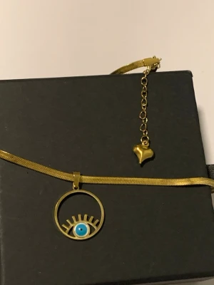 Guldigt halsband med ögonberlock - Unikt halsband i guldfärgad metall med en rund berlock som föreställer ett öga med blå detalj och fransar. Kedjan har en liten hjärtformad detalj vid förlängningen. Perfekt för dig som gillar statement-smycken och trendiga accessoarer.