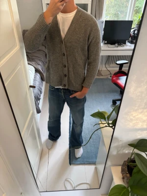 Grå stickad kofta med knappar - Mysig grå stickad kofta med v-ringning och fyra knappar framtill. Koftan har två fickor och ribbade muddar vid ärmslut och nederkant. Perfekt att slänga över en t-shirt för en avslappnad look.