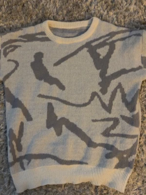 SEGNO Tröja - Säljer en beige stickad tröja med abstrakt grått mönster. Tröjan har rund halsringning, ribbade muddar och nederkant. Perfekt för dig som gillar en unik och modern stil. Materialet känns mjukt och bekvämt.
