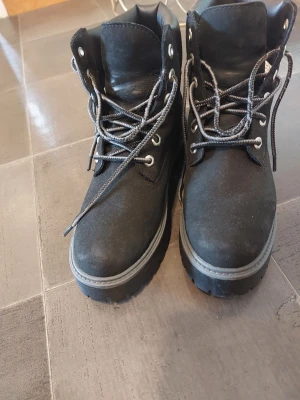 Svarta kängor från Timberland - Säljer ett par svarta kängor från Timberland med grov sula och vadderad kant. Kängorna har snörning med gråa skosnören och metallhål. Tillverkade i mocka och läder, perfekta för höst och vinter.