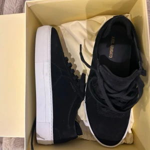 Oanvända Svarta sneakers från Axel Arigato - Stilrena svarta sneakers från Axel Arigato med ovandel i mocka och klassisk vit sula. Skorna har svarta skosnören och diskret logga i guld på sidan. 