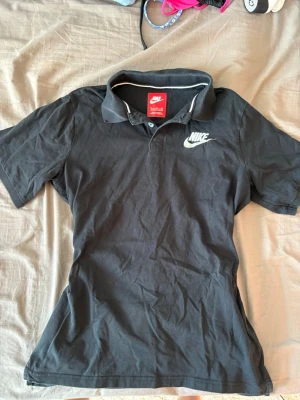 Svart pikétröja från Nike - Klassisk svart pikétröja från Nike med vit logga på bröstet och krage med diskret vit kant. Tröjan har korta ärmar och normal passform, perfekt för en sportig och avslappnad stil.