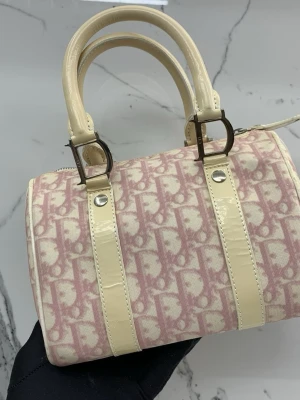 Rosa och beige monogram handväska - Snygg handväska i boxig modell med rosa och beige monogrammönster. Väskan har två korta handtag i glansigt beige material och detaljer i metall. Perfekt för dig som gillar en trendig och klassisk look.   ena handtaget är av men går att fixa, säljs i befintligt skick