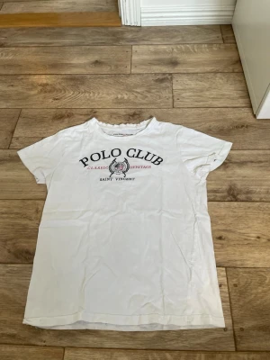 Vit Polo Club t-shirt med tryck - Vit t-shirt från Polo Club med klassiskt tryck i svart och rött på bröstet. Rund halsringning och korta ärmar. Tillverkad i mjuk bomull för en skön känsla. Perfekt till jeans eller shorts för en avslappnad look. Storlek 150 