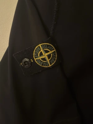 Svart jacka från Stone Island Junior - Säljer en svart jacka från Stone Island Junior med den klassiska logotypen på ärmen. Jackan har en stilren design och är tillverkad i ett slätt material som passar perfekt till streetwear. Perfekt för dig som gillar exklusiva märken och snygg stil.