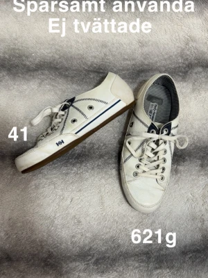 Vita sneakers från Helly Hansen - Säljer ett par vita sneakers från Helly Hansen med marinblå detaljer och sicksack-sömmar. Skorna har snörning, randigt innerfoder och slitstark gummisula. Klassisk låg modell som funkar till det mesta.🌸 Sparsamt använda. Ej tvättade. Överdelen är gjord av läder. Storlek 41 