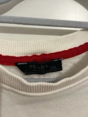 Vit långärmad tröja från Prada - Säljer en vit långärmad tröja från Prada med rund halsringning och röda detaljer. Tröjan har en broderad Prada-logga på bröstet och en ikonisk trekantig patch på ärmen. Tillverkad i mjukt tyg som känns skönt mot huden. Går för XL den är mindre än vad det står 