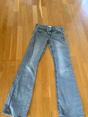 Grå bootcut jeans med fickor - Säljer ett par grå bootcut jeans med klassisk femficksdesign och knappgylf. Jeansen har lätt slitna detaljer framtill och bakfickor med lock. 