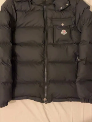 Svart dunjacka från Moncler - Svart dunjacka från Moncler med avtagbar huva, dragkedja framtill och en bröstficka med knapp och Moncler-logga. Jackan har två sidofickor med dragkedja och en klassisk puffer-design som håller dig varm under kalla dagar.