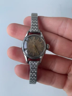 Vintage Omega klocka i silver - Klassisk vintage Omega armbandsur med rund urtavla och stilrent silverfärgat metallarmband. Urtavlan har en unik patina och tydliga indexmarkeringar. Perfekt accessoar för dig som gillar retrostil och tidlös design. El liten repa i glaset men går att fixa. Behövs nog ett nytt batteri. Tickar ibland. Men en riktigt fin klocka!