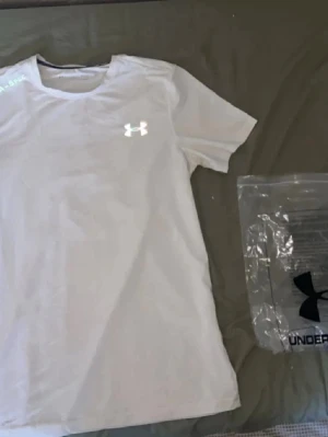 Vit Under Armour t-shirt - Storlek M, Snygg vit t-shirt från Under Armour med UA-logga på bröstet och texten UA-SPEC vid axeln. Klassisk rund hals och korta ärmar. Perfekt för dig som gillar sportig och clean stil.