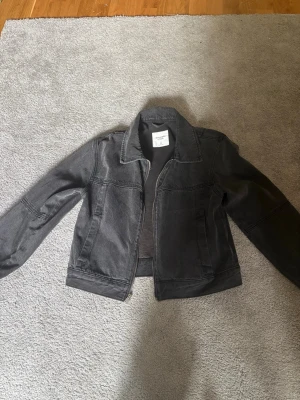 Mörkgrå jeansjacka med rak passform - Snygg mörkgrå jeansjacka med klassisk krage och raka sömmar. Jackan har två sidofickor och stängs med dragkedja framtill. Perfekt för dig som gillar en enkel men trendig look. Passar till det mesta och är lätt att matcha.