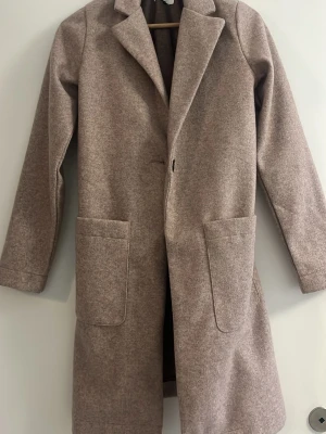 Beige ullkappa med stora fickor - Stilren beige kappa i ullmix med klassisk krage och stora utanpåfickor. Rak passform och enkel knäppning framtill. Ofodrad passar till vår eller höst
