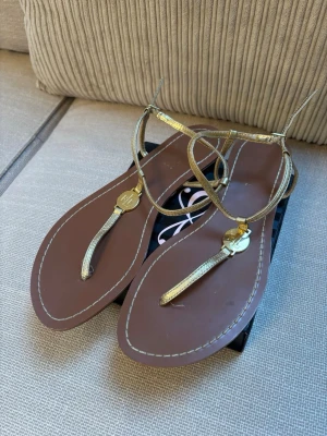 Guldiga sandaler från Ralph Lauren  - Snygga sandaler från Ralph Lauren med tunna, guldiga remmar och bruna sulor. Sandalerna har en elegant T-rem med metallisk logodetalj framtill och kontrastsömmar runt sulan. Perfekta för sommarens alla outfits och ger en lyxig touch till din stil. Köparen betalar frakten.