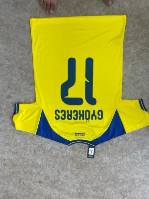 Gul Sverige fotbollströja nr 17 - Säljer en gul fotbollströja från Sverige med blå detaljer och nummer 17 på ryggen. Tröjan har trycket 'GYOKERES' och svenska flaggan på framsidan. Klassisk sportdesign med rund hals och korta ärmar, perfekt för match eller träning.