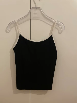 Svart linnetopp från BikBok - Snygg svart linnetopp från BikBok med tunna axelband och vita kanter. Enkel och stilren design som passar perfekt till jeans eller kjol. Tillverkad i mjukt material för en bekväm känsla.