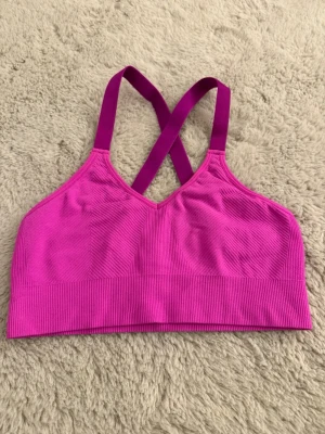 Rosa sport-bh - Snygg sport-bh i stark rosa färg med ribbad struktur och bred resår nedtill. Bh:n har korsade axelband i ryggen för extra stöd och en sportig look. Materialet är stretchigt och mjukt, perfekt för träning eller chill. 