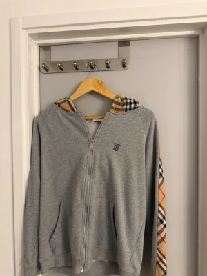 Grå Burberry hoodie med dragkedja - Snygg grå hoodie från Burberry med dragkedja framtill och klassiskt rutigt mönster i huvan och längs ena ärmen. Har två fickor fram och en liten broderad Burberry-logga på bröstet. Perfekt för en chill och trendig look.
