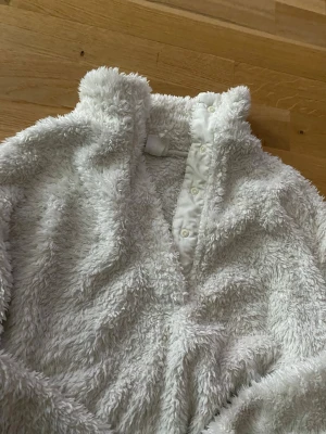 Vitt fluffigt loungewear från H&M - Supermjuk och fluffig vit loungewear-tröja från H&M med hög krage och knappar framtill. Perfekt för mysiga hemmakvällar. Tillverkad i ett lurvigt material som känns som en kram. Enkel och stilren design.