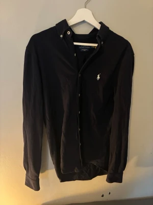 Mörkblå skjorta från Ralph Lauren - Klassisk mörkblå skjorta från Ralph Lauren med vit broderad logga på bröstet. Skjortan har långa ärmar, knappar framtill och en tidlös design som passar till det mesta. Tillverkad i mjuk bomull för skön känsla. - vid snabba köp kan priset sänkas🔥