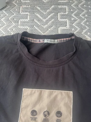 Svart Burberry t-shirt med beige patch - Snygg svart t-shirt från Burberry med rund hals och en beige patch på bröstet med Burberry-logga och tryck. Klassisk passform och diskret Burberry-mönster på insidan av kragen. Perfekt för en clean och stilren look.