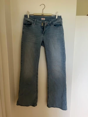 Ljusblå bootcut jeans med brodyr - Säljer ett par ljusblå bootcut jeans med dubbla knappar och broderade detaljer i rött på bakfickorna. Jeansen är tillverkade i denim med lätt tvättad look. Perfekta för en avslappnad och trendig stil.