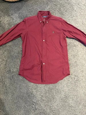 Vinröd skjorta från Polo Ralph Lauren - Klassisk vinröd skjorta från Polo Ralph Lauren med button-down krage och broderad logga på bröstet. Skjortan har lång ärm, knappar framtill och är tillverkad i bomull. Perfekt för en stilren och avslappnad look. Riktigt bra skick, endast använd 2 gånger men är lite små skrynklig men det är bara att stryka skjortan så är den så gott som ny.