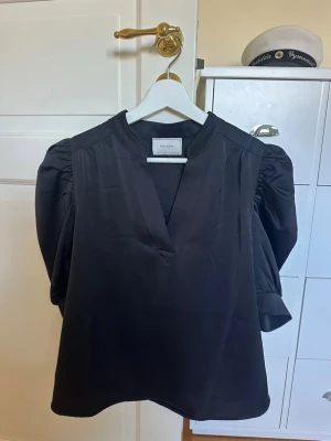 Svart blus med puffärm och v-ringning - Stilren svart blus med v-ringning och puffiga långärmar. Blusen har en rak passform och lätt glansigt material som ger en elegant känsla. Perfekt att matcha med både jeans och kjol för en trendig look.
